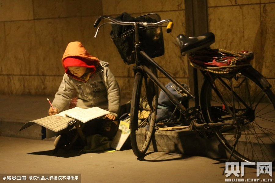 鄭州中原路京廣鐵路大橋下涵洞的馬路牙子邊，一位戴著紅色辣椒帽的小男孩趴在一個小馬扎上，上面鋪著一張車墊，在微弱的燈光下寫作業(yè)